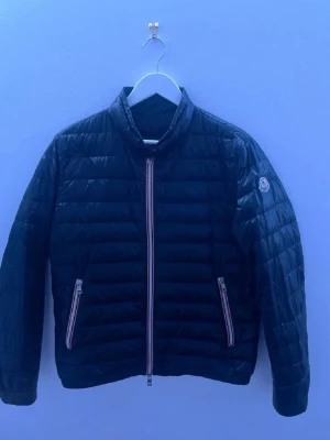Svart dunjacka från Moncler - Snygg svart dunjacka från Moncler med quiltad design och ståkrage. Jackan har två framfickor med dragkedja och Moncler-logga på vänster ärm. Dragkedjorna har rödvita detaljer som ger en sportig touch. Perfekt för kalla vinterdagar.