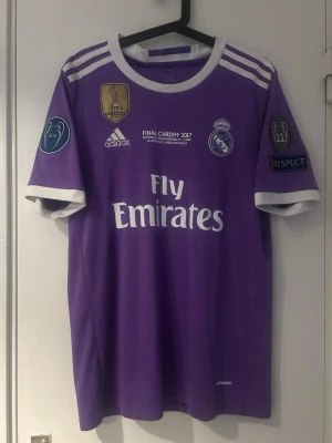 Lila Real Madrid Ronaldo tröja - Säljer en lila Real Madrid matchtröja från Adidas med Ronaldo 7 på ryggen. Tröjan har vita detaljer, Champions League och FIFA-märken samt Fly Emirates-tryck på bröstet. Perfekt för dig som älskar fotboll och vill ha en ikonisk supportertröja.