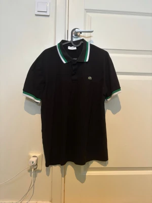 Svart pikétröja från Lacoste - Snygg svart pikétröja från Lacoste med klassisk krage och gröna samt vita ränder på krage och ärmslut. I princip helt ny, inga tecken på användning. Pris går att diskuteras. Kan skickas bild på längd och bredd vid intresse. 