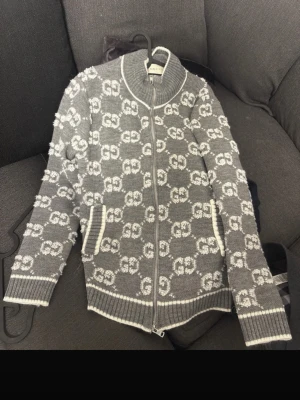 Grå ziptröja med GG-mönster från Gucci - Snygg grå ziptröja från Gucci med vitt GG-monogram över hela tröjan. Tröjan har ribbade muddar, hög krage och dragkedja framtill. Tillverkad i mjukt stickat material och har vita detaljer vid fickor, ärmslut och nederkant.