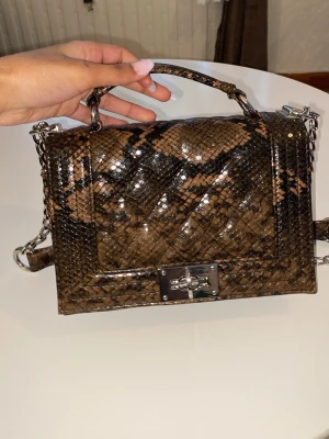 Brun axelväska med ormmönster - Snygg brun axelväska med ormmönster och glansig finish. Väskan har silverfärgad kedjerem, ett handtag upptill och ett metallspänne framtill. Perfekt för dig som vill sticka ut med en trendig accessoar. Materialet ser ut som konstläder.