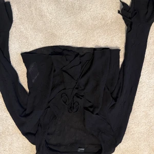 Svart topp från Gina  - Snygg svart topp med långa ärmar och knytning framtill. Toppen har en lätt skrynklig struktur och är perfekt för dig som gillar en stilren look med det lilla extra. Passar dig som vill ha en enkel men trendig topp.