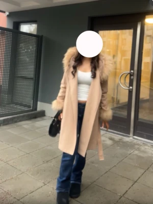 Beige kappa med fuskpälsdetaljer - Stilren beige kappa med stora fuskpälsdetaljer vid krage och ärmslut. Kappan har knytskärp i midjan och rak passform som ger en snygg siluett. Perfekt för kyligare dagar och enkel att matcha med olika outfits.