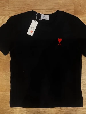 Svart t-shirt från Ami Paris - Svart t-shirt från Ami Paris med röd broderad logga i form av ett hjärta och bokstaven A på bröstet. Klassisk rund halsringning och korta ärmar. Tillverkad i mjuk bomull för en skön känsla och stilren look.