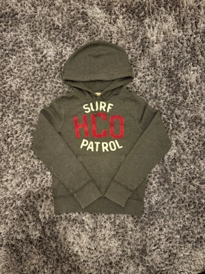 Grå hollister hoodie - Mörkgrå hoodie från Hollister med stor huva och dragsko. Framtill står det 'SURF HCO PATROL' i rött och vitt tryck. Klassisk känguruficka och mjukt material, perfekt för chill dagar. Oversized känsla och skön passform.
