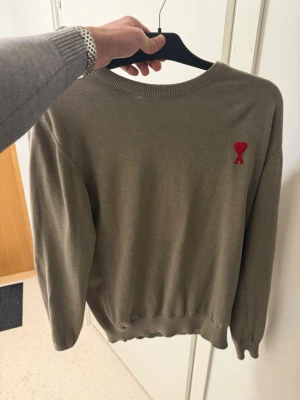 Beige Ami Paris  - Snygg beige tröja från Ami Paris med den ikoniska röda hjärta-A-loggan broderad på bröstet. Storlek L men sitter som M! Tröjan har rund halsringning, ribbade muddar och är tillverkad i mjukt material som känns skönt mot huden. Perfekt för en clean och stilren look.