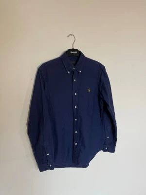 Marinblå skjorta från Polo Ralph Lauren - Ralph Lauren skjorta, Marinblå  Storlek M slim fit  Mycket fint skick   Kolla gärna in mina plagg så får ni bundles rabatt Klassisk marinblå skjorta från Polo Ralph Lauren med button-down krage och broderad gul logga på bröstet. Skjortan har långa ärmar och vita knappar framtill samt vid ärmsluten. Perfekt till en clean och stilren look.