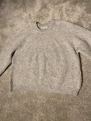 Grå stickad tröja - Mysig grå stickad tröja med rund halsringning och ribbstickat mönster. Tröjan har långa ärmar och en avslappnad passform. Perfekt för lager på lager och enkel att matcha med olika stilar.