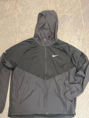 Svart vindjacka från Nike med huva - Snygg svart vindjacka från Nike med dragkedja framtill och huva. Jackan har två sidofickor med dragkedja och en vit Nike-logga på bröstet. Tillverkad i lätt och tåligt material, perfekt för blåsiga dagar.