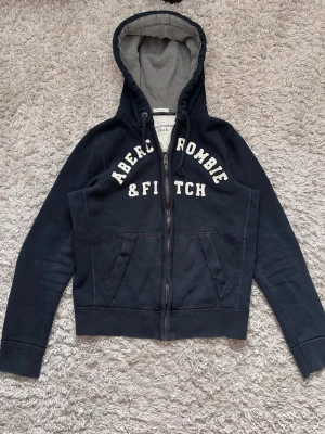 Mörkblå hoodie från Abercrombie & Fitch - Snygg mörkblå hoodie med dragkedja från Abercrombie & Fitch. Stor vit logga på bröstet, två fickor fram och ribbade muddar. Insidan av huvan är grå och mjuk för extra komfort. Perfekt för en avslappnad och trendig look.
