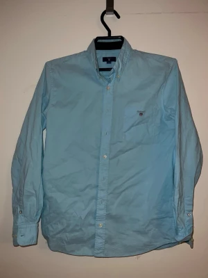 Ljusblå skjorta från Gant - DEN STRYKS INNAN FRAKT! Klassisk ljusblå skjorta från Gant med button-down krage och bröstficka med broderad logga. Skjortan har långa ärmar och vita knappar framtill. Perfekt till en clean och stilren look. Passar mer åt grisch hållet men funkar till alla sorts tillfällen