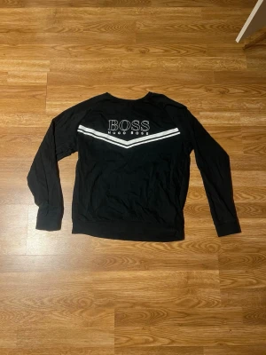 Svart sweatshirt från Hugo Boss - Svart sweatshirt med lång ärm från Hugo Boss. Tröjan har vit logga och två vita ränder över bröstet. Klassisk rund halsringning och ribbade muddar vid ärmslut och nederkant. Perfekt för en avslappnad och stilren look. Perfekt för en stilren look och passar till de mesta 