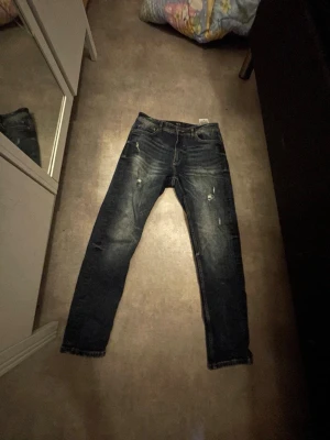 Mörkblå slitna jeans med  detaljer - Säljer ett par mörkblå jeans med slitningar och  detaljer på både fram- och baksida. Jeansen har fem fickor, knappgylf och en klassisk passform. Materialet är denim och de har en cool, avslappnad look.