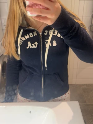 Mörkblå hoodie Abercrombie & Fitch - Mörkblå zip-hoodie från Abercrombie & Fitch med vita snören och broderad logga på bröstet. Hoodien har fickor framtill och en mjuk, ljusgrå insida i huvan. Perfekt för en avslappnad och cool stil.