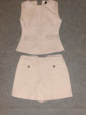 Ljusrosa bouclé set med topp & shorts - Säljer ett ljusrosa bouclé set med ärmlös topp och matchande shorts. Toppen har figurnära passform och dekorativa fickor framtill. Shortsen har två fickor med guldfärgade knappar. Materialet är strukturerat och ger en lyxig känsla.