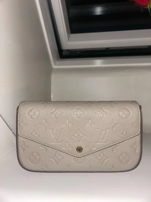 Beige Louis Vuitton kuvertväska - Säljer en beige kuvertväska från Louis Vuitton i präglat läder med det klassiska monogrammönstret. Väskan har en elegant, avlång form och stängs med en guldfärgad knapp framtill. Perfekt accessoar för dig som gillar lyxiga detaljer och stilren design.