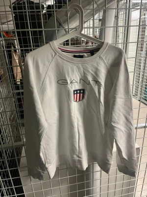 Vit sweatshirt från GANT med logga - Vit sweatshirt från GANT med broderad logga och sköld med stjärnor och ränder på bröstet. Tröjan har rund halsringning med röd och marinblå rand på insidan och långa ärmar. Perfekt för en clean och sportig stil.