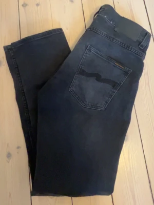 Svarta/mörkgråa jeans grim trim från Nudie Jeans - Säljer ett par svarta jeans från Nudie Jeans, grim trim med klassisk femficksmodell och diskret logga på bakfickan. Jeansen har en smal passform och är tillverkade i mjukt denimtyg. Perfekta för en stilren och avslappnad look.