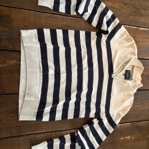 Randig tröja från Bondelid  - Säljer en långärmad tröja från Bondelid med breda marinblå och vita ränder. Tröjan har krage och kort dragkedja vid halsen för en sportig look. Tillverkad i mjukt stickat material som känns skönt mot huden. Perfekt för dig som gillar klassisk stil.