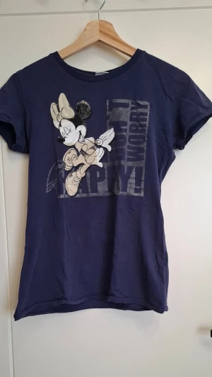 Blå Disney t-shirt med Minnie Mouse - Mörkblå t-shirt från Disney med tryck av Minnie Mouse och texten 'Don't Worry Be Happy!' på framsidan. T-shirten har korta ärmar och rund hals. Perfekt för dig som gillar klassiska Disney-karaktärer och vill ha en avslappnad stil.