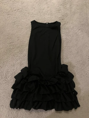 Svart volangklänning med shorts - Supersnygg svart ribbad klänning med volanger nertill och inbyggda shorts under kjolen. Ärmlös modell med rund halsringning och dragkedja i ryggen. Perfekt för dig som vill ha både stil och komfort. Materialet känns mjukt och följsamt. Den har också slitsar på bägge sidor av kjolen som gör att shortsen under syns…