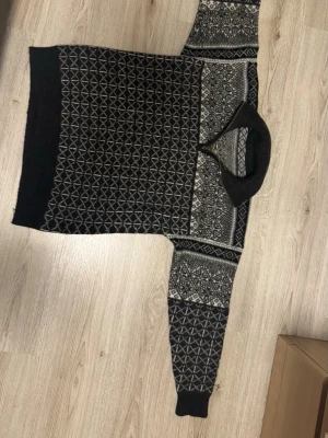 Svartvit stickad polotröja med mönster - Snygg svart stickad polotröja med vita geometriska och nordiska mönster. Tröjan har hög krage och ribbade muddar. Perfekt för kalla dagar när du vill vara både varm och stilren. 100% Ull