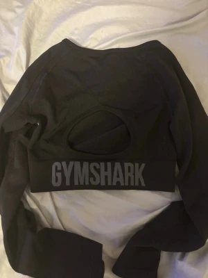 Svart croppad topp från Gymshark - Snygg svart croppad topp från Gymshark med lång ärm. Toppen har ett diskret Gymshark-tryck framtill och ett större logotyptryck bak. Tillverkad i stretchigt syntetmaterial som passar perfekt till träning. Använd 1 gång.