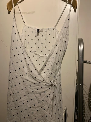 Vit omlottklänning med prickar - Söt vit omlottklänning med svarta prickar och smala axelband. Klänningen har knyt i sidan och en djup v-ringning framtill. Perfekt för varma dagar och ger en fräsch och trendig look.