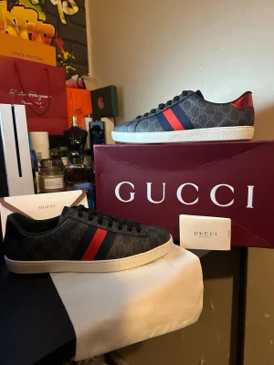 Gucci Ace sneakers med röd/blå rand - Snygga Gucci Ace sneakers i svart läder med mörkgrå GG-monogram, röd och blå sidrand samt metallic-detaljer i rött och blått på hälen. Klassisk låg modell med vit sula och svarta skosnören. Perfekta för dig som vill ha en lyxig och trendig look.