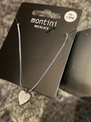 Silverfärgat hjärthalsband Montini - Snyggt halsband från Montini i silverfärg med ett hjärtformat hänge. Kedjan är tunn och stilren, perfekt för att ge en söt touch till din outfit. Hjärtat har en blank finish och halsbandet är enkelt att matcha med andra smycken.