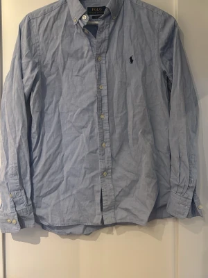 Blå skjorta från Polo Ralph Lauren - Klassisk ljusblå skjorta från Polo Ralph Lauren. Skjortan är i finfint skick om man bortser ifrån det extremt lilla hålet på nedre baksidan (se bild 3). Skjortan är ca 165 stor. Skjortan är perfekt till sommaren och är sparsamt använd. Hör av er vid frågor o funderingar 🍾