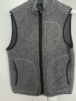 J.lindeberg fleece väst - Mysig grå väst i fleece fårn j.lindeberg med hel dragkedja framtill, hög krage och svarta kantband. Västen har tre fickor med dragkedja och en stilren, enkel design. Perfekt för lager-på-lager och håller dig varm under kyliga dagar. Köpt på j.lindeberh butik för 2299 kr