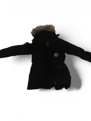 Svart parkas från Canada Goose - Svart parkasjacka från Canada Goose med klassisk patch på bröstet, flera stora fickor och huva med fuskpäls. Jackan har rak passform och dragkedja framtill. Perfekt för kalla dagar när du vill hålla stilen och värmen. Det ända dåliga med jackan är att dragkedjan inte funka 