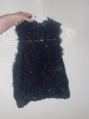 Svart spetsig ärmlös topp med dragkedja - Secondhandat Snyggt svart ärmlös topp med spetsdetaljer och en cool dragkedja i ryggen. Toppen har en unik textur och är perfekt för att sticka ut. Passar dig som gillar trendiga och lite edgy plagg.