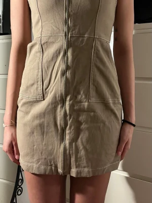 Beige kort klänning med dragkedja - Snygg beige kort klänning i bomull med markerade sömmar och dragkedja framtill. Klänningen har breda axelband och två stora fickor framtill, perfekt för en trendig och avslappnad look.