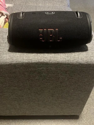 JBL svart trådlös högtalare - Kraftfull trådlös högtalare från JBL i svart med robust cylinderformad design. Högtalaren har ett slitstarkt tygmaterial och gummerade ändar för extra skydd. JBL-loggan syns tydligt på framsidan och högtalaren har smidiga knappar på ovansidan för enkel styrning.