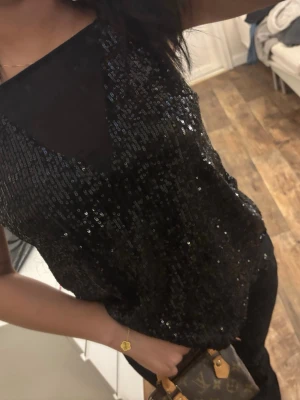 Svart glittrig topp med meshdetalj - Snygg svart topp med glittriga paljetter och en transparent meshdetalj upptill. Perfekt för dig som vill sticka ut och glänsa. Toppens passform är normal och den är ärmlös, vilket ger en cool och trendig look.
