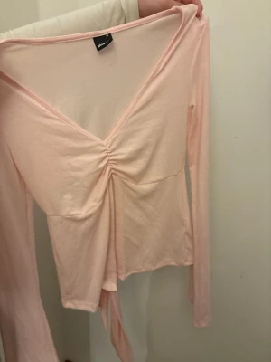 Ljusrosa omlottopp med v-ringning - Supersöt ljusrosa omlottopp från Gina Tricot med v-ringning och rynkdetalj framtill. Långärmad och i mjukt, stretchigt material som känns skönt mot huden. Perfekt att styla med jeans eller kjol för en trendig look.