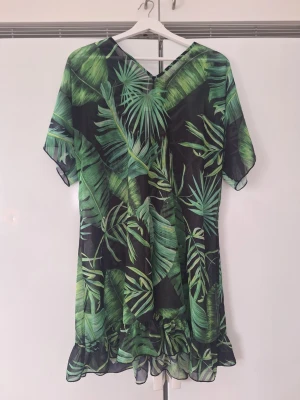 Grön strandklänning med palmblad - Snygg strandklänning i transparent material med tropiskt palmbladsmönster i olika gröna nyanser. Klänningen har korta ärmar, v-ringning och volangkant nertill. Perfekt att dra över bikinin på stranden. Knytning i nacken för extra detalj. Passar de flesta.