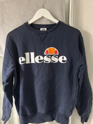 Mörkblå sweatshirt från Ellesse - Mörkblå klassik sweatshirt från Ellesse med stort  logotryck på bröstet. Den är liten i storleken så tänk på det ifall du har långa armar. Legat nervikt i garderoben där av att den ser skrynklig ut på bilden men tröjan är helt ren och utan märken 