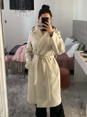 Beige lång kappa med huva och knytskärp - Säljer en stilren beige kappa med huva och knytskärp i midjan. Kappan har en mjuk och varm känsla, perfekt för kyliga dagar. Den är lång och har en klassisk omlott-design som ger en snygg siluett. Passar dig som vill ha en enkel men trendig look.
