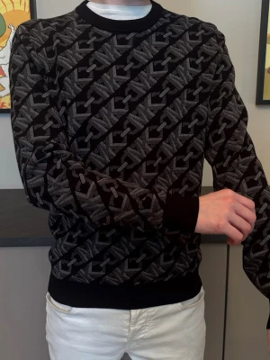 Stickad Michael Kors Tröja - Säljer en svart stickad tröja med grått diagonal logomönster över hela plagget. Tröjan är otroligt töjbar, har rund halsringning, ribbade muddar vid ärmslut och nederkant samt en slim fit passform. Tveka inte, ställ frågor och kom med pris förslag! 