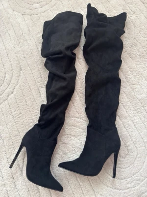 Svarta overknee stövlar med klack - Säljer ett par svarta overknee stövlar. De har hög smal klack, spetsig tå och är gjorda i mjukt mockaimitationsmaterial. Stövlarna är slouchy och sitter snyggt över knät för en trendig look.