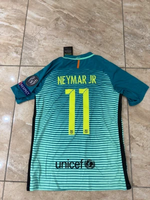Barcelona Neymar JR 2016/17 matchtröja - Säljer en Barcelona matchtröja från Nike med Neymar JR och nummer 11 på ryggen. Tröjan är turkos med svarta och ljusblå ränder, har korta ärmar och klubbmärke samt sponsortryck på framsidan. Perfekt för fotbollsfans som vill sticka ut.