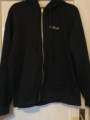 Svart zip hoodie från Replay - Säljer en svart hoodie från Replay med dragkedja framtill och broderad logga på bröstet. Hoodien har huva med dragsko och en stor magficka. Perfekt för dig som gillar stilrena och klassiska plagg. Materialet känns mjukt och bekvämt.