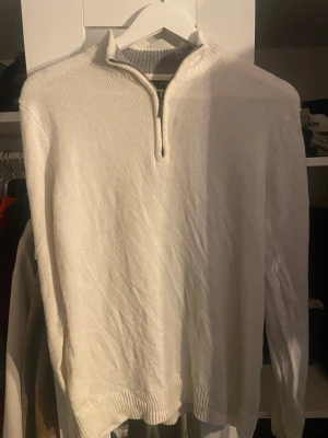 Vit stickad tröja med halv dragkedja - Snygg vit stickad tröja med halv dragkedja och ståkrage. Tröjan har långa ärmar och ribbade muddar vid ärmslut och nederkant. Perfekt för en stilren och avslappnad look.