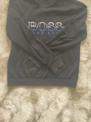 Svart sweatshirt från Hugo Boss - Snygg svart sweatshirt från Hugo Boss med stor vit och blå logga på bröstet. Klassisk rund halsringning och ribbade muddar vid ärmslut och nederkant. Perfekt för en avslappnad och stilren look.