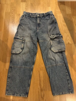 Grå cargobyxor i denim med stora fickor - Size 38. Säljer ett par grå cargobyxor i tvättad denim med bred passform och stora fickor på sidorna. Byxorna har klassisk cargostil med flera fickor och markerade sömmar över benen. Perfekta för en avslappnad streetwear-look.
