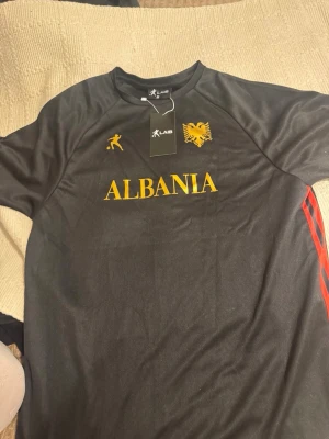 Svart Albanien fotbollströja LAIB - Snygg svart fotbollströja från LAIB med gult Albanien-tryck, örnlogga och fotbollsspelare på bröstet. Tröjan har röda ränder på sidan och är gjord i ett lätt och sportigt material som passar perfekt till träning eller match.