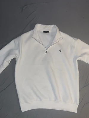 Vit quarter zip från Polo Ralph Lauren  - Snygg vit quarter zip sweatshirt från Polo Ralph Lauren med klassisk broderad logga på bröstet. Tröjan har hög krage, dragkedja framtill och ribbade muddar vid ärmslut och nederkant. Perfekt för en clean och stilren look.